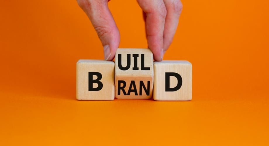 Hand arranging wooden blocks spelling out 'BUILD BRAND' against an orange background.